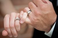 A close-up of a bride and groom's left hands, where the bride's engagement ring set and the groom's wedding band are front and center.